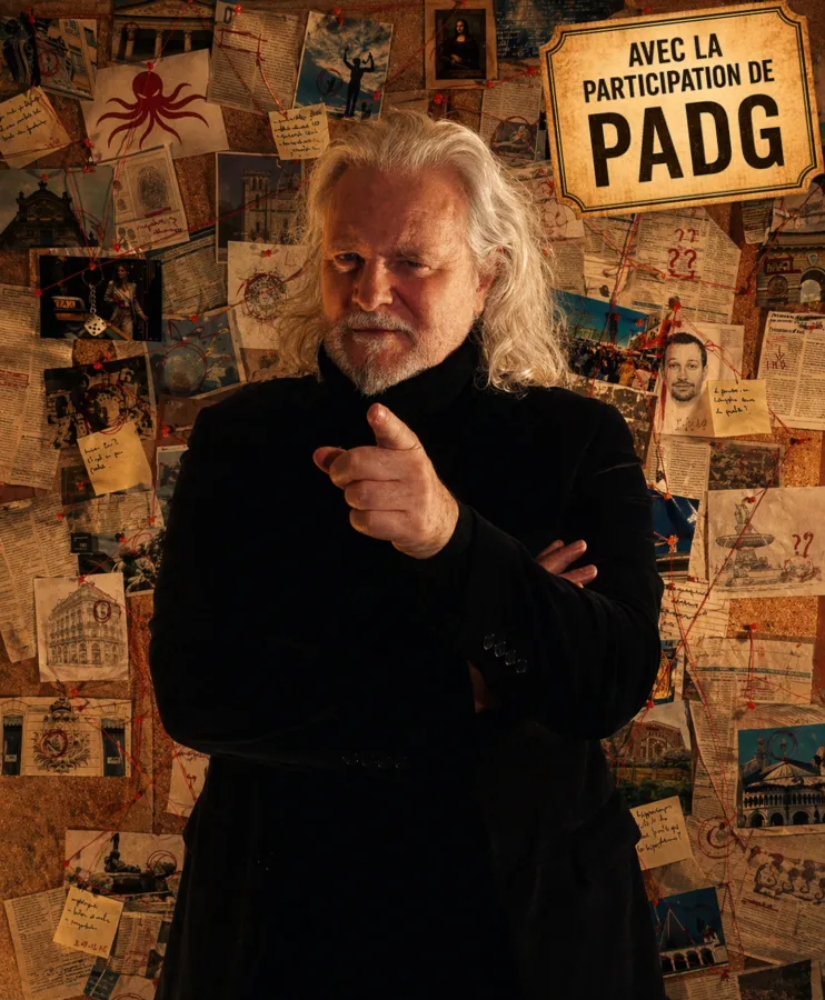 Man with long white hair wearing a black suit, pointing at the camera, standing in front of a wall covered with pinned photos, drawings and documents, with a sign reading 'AVEC LA PARTICIPATION DE PADG' in Bordeaux.