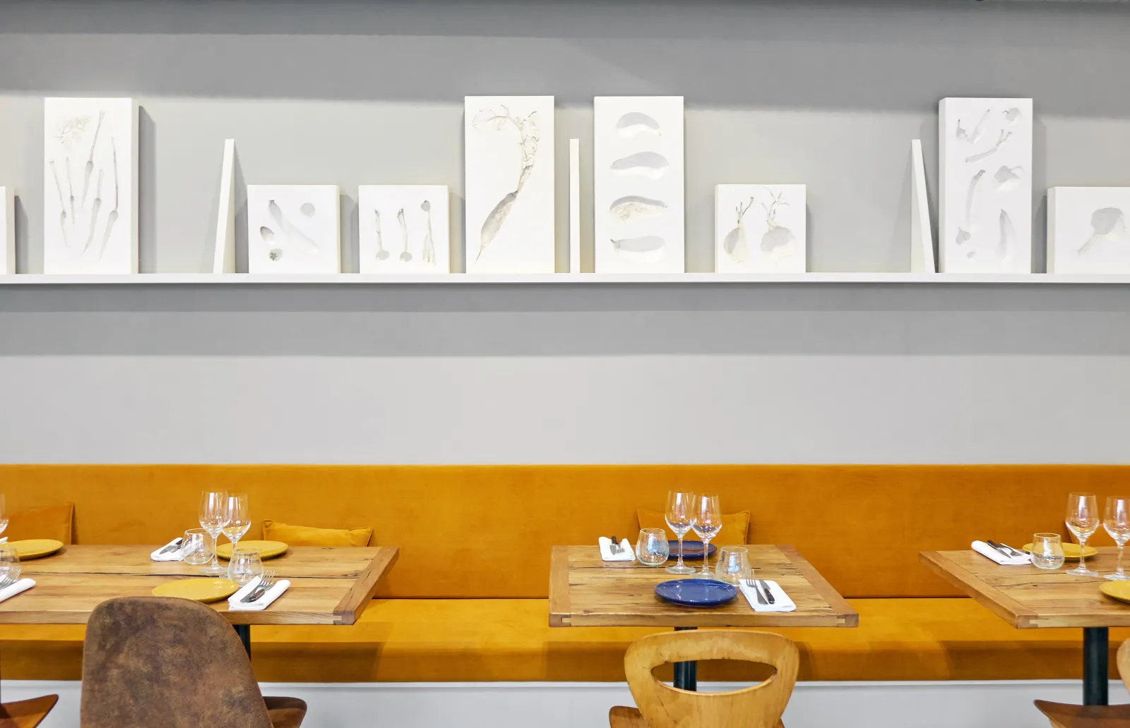 Modern restaurant interior featuring a vibrant yellow banquette, wooden tables with place settings, glasses, and a white floating shelf displaying minimalist botanical sketches