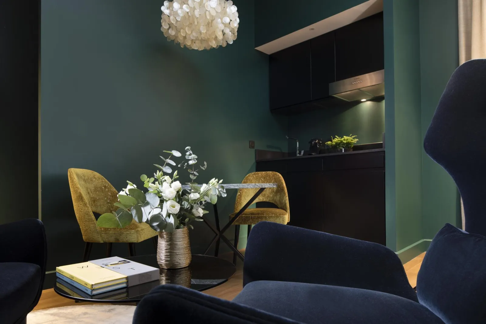 Elegant reception area of the Hôtel Cardinal Bordeaux featuring sage green walls, golden-yellow chairs, marble table, white floral arrangement, and modern shell-like pendant light.