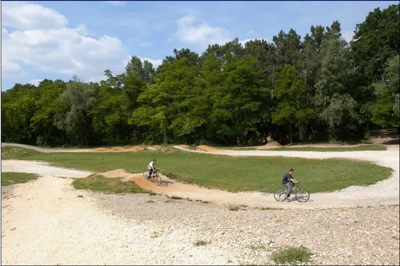 Two cyclists riding on a trial bike course in Bois du Burck near Bordeaux, featuring dirt jumps and berms surrounded by lush forest.