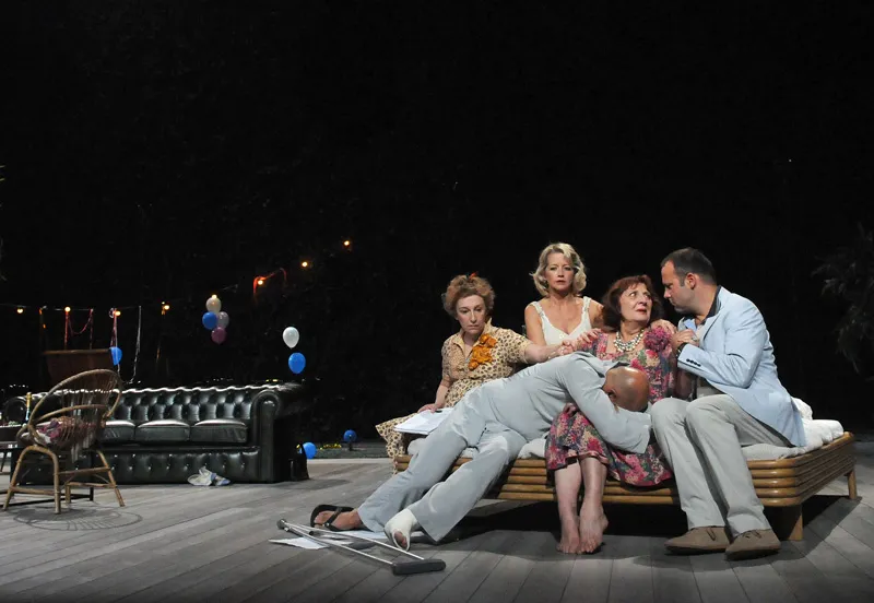 Theater scene at the National Theater Bordeaux in Aquitaine featuring five actors in 1950s-style costumes seated on vintage couches, set in a stylized interior with period furniture and colored balloons.