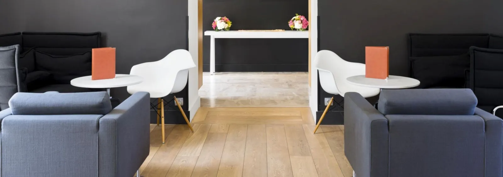 Modern interior of Saint-James in Bordeaux featuring two symmetrical white chairs, gray sofas, white side tables with orange books, light oak flooring and a floral-decorated reception desk in the background.