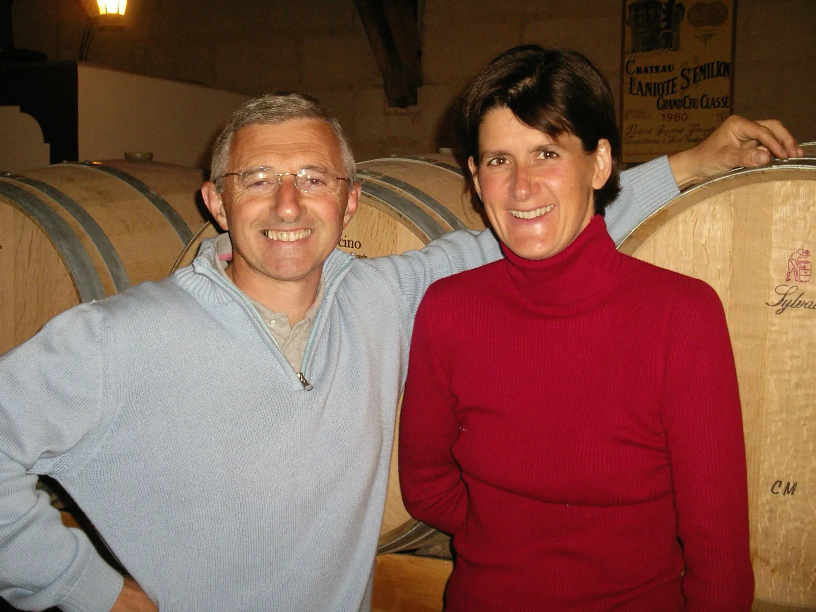 Two smiling people posing together in a wine cellar at Château Laniote in Bordeaux, surrounded by oak barrels.