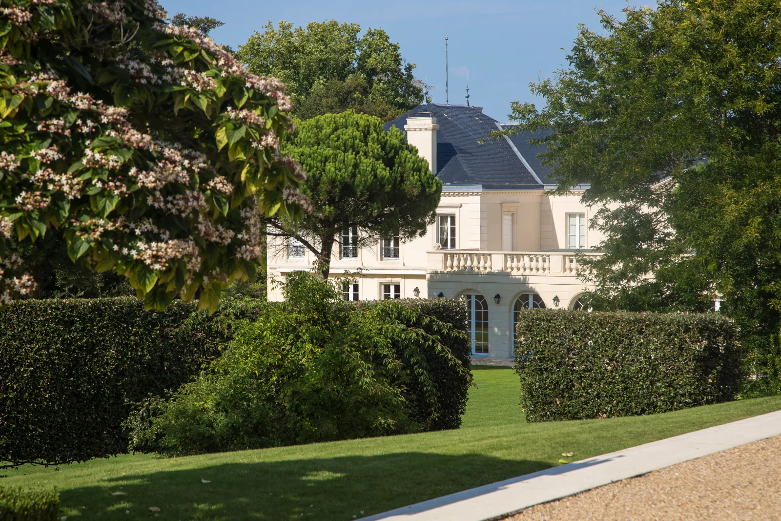View of Château Malartic-Lagravière in Bordeaux, white facade with slate roof surrounded by formal French gardens with trimmed hedges and blooming lilac flowers