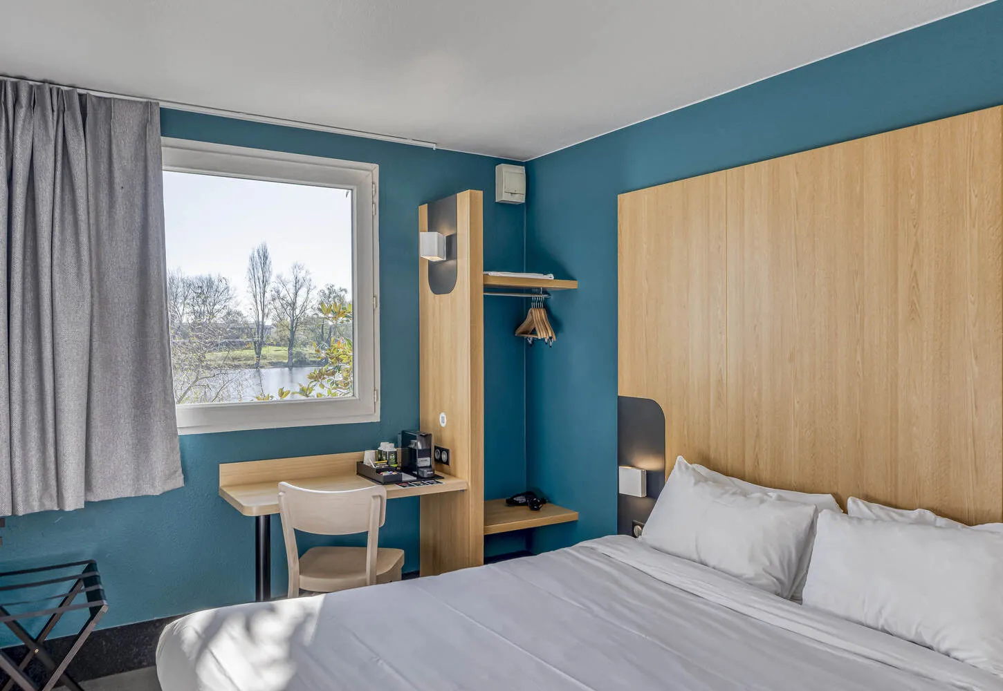 Modern hotel room with teal walls, white bedding, natural wood furniture, desk with chair, and scenic countryside view through the window