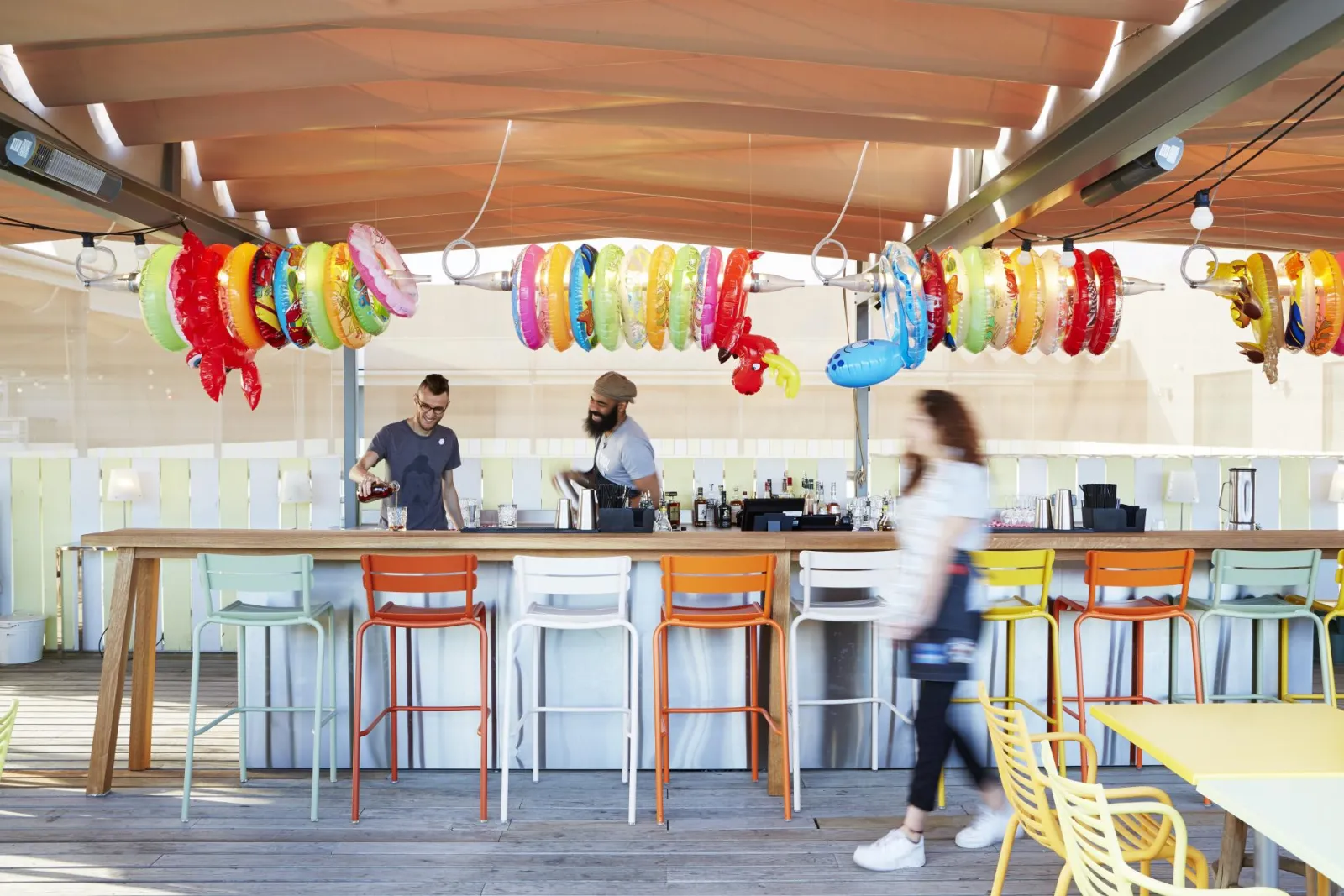 Modern bar at Mama Shelter in Bordeaux featuring wooden counter, colorful bar stools, staff members working, and festive hanging bracelet decorations on the ceiling.