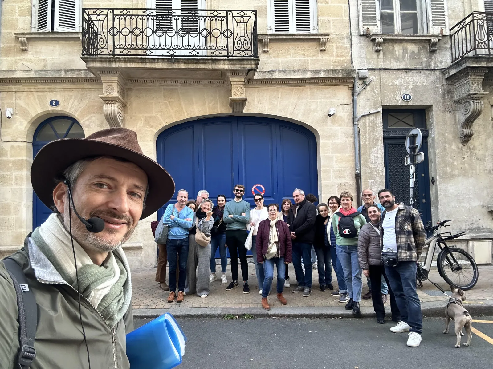 A tour guide wearing a brown hat smiles at the camera during a guided tour in front of a beautiful Bordeaux facade with a large blue door, with a group of tourists posing in the background.