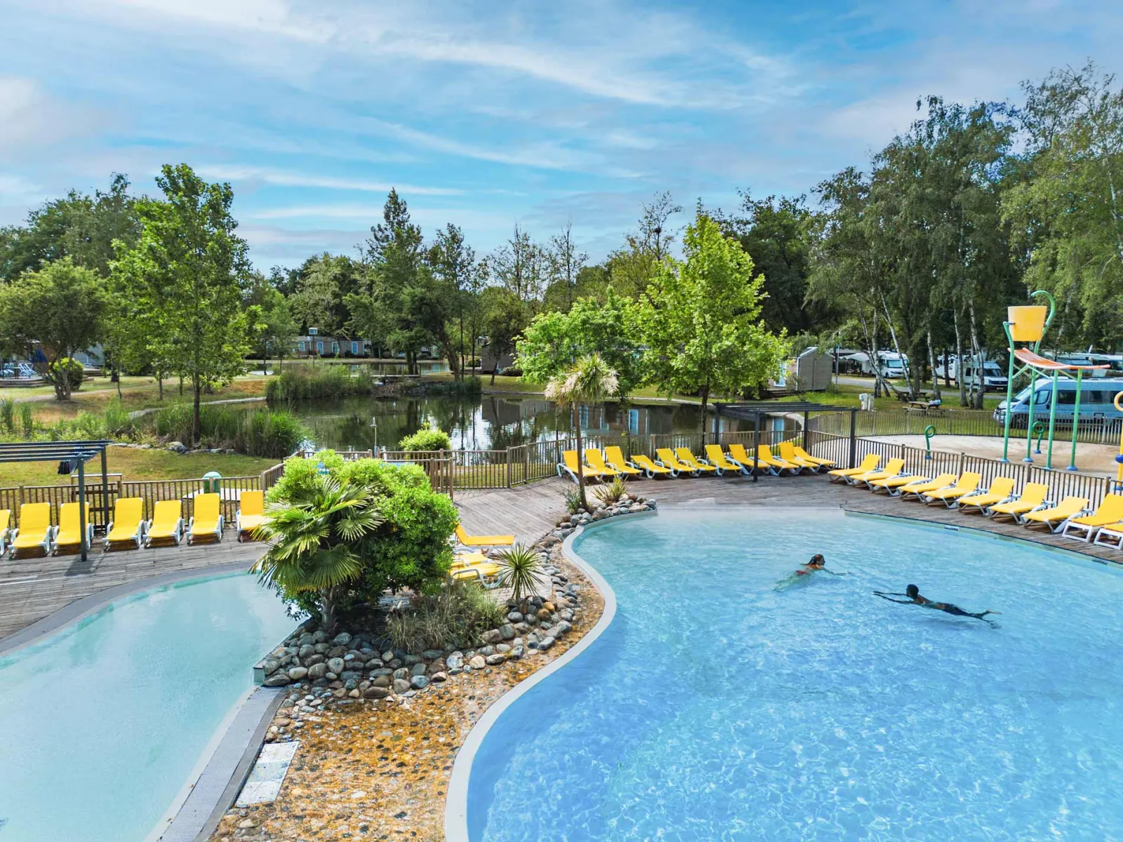 Outdoor swimming pool with yellow lounge chairs, shallow pools, lush greenery and lake in the background at Yelloh! Village Bordeaux Lac