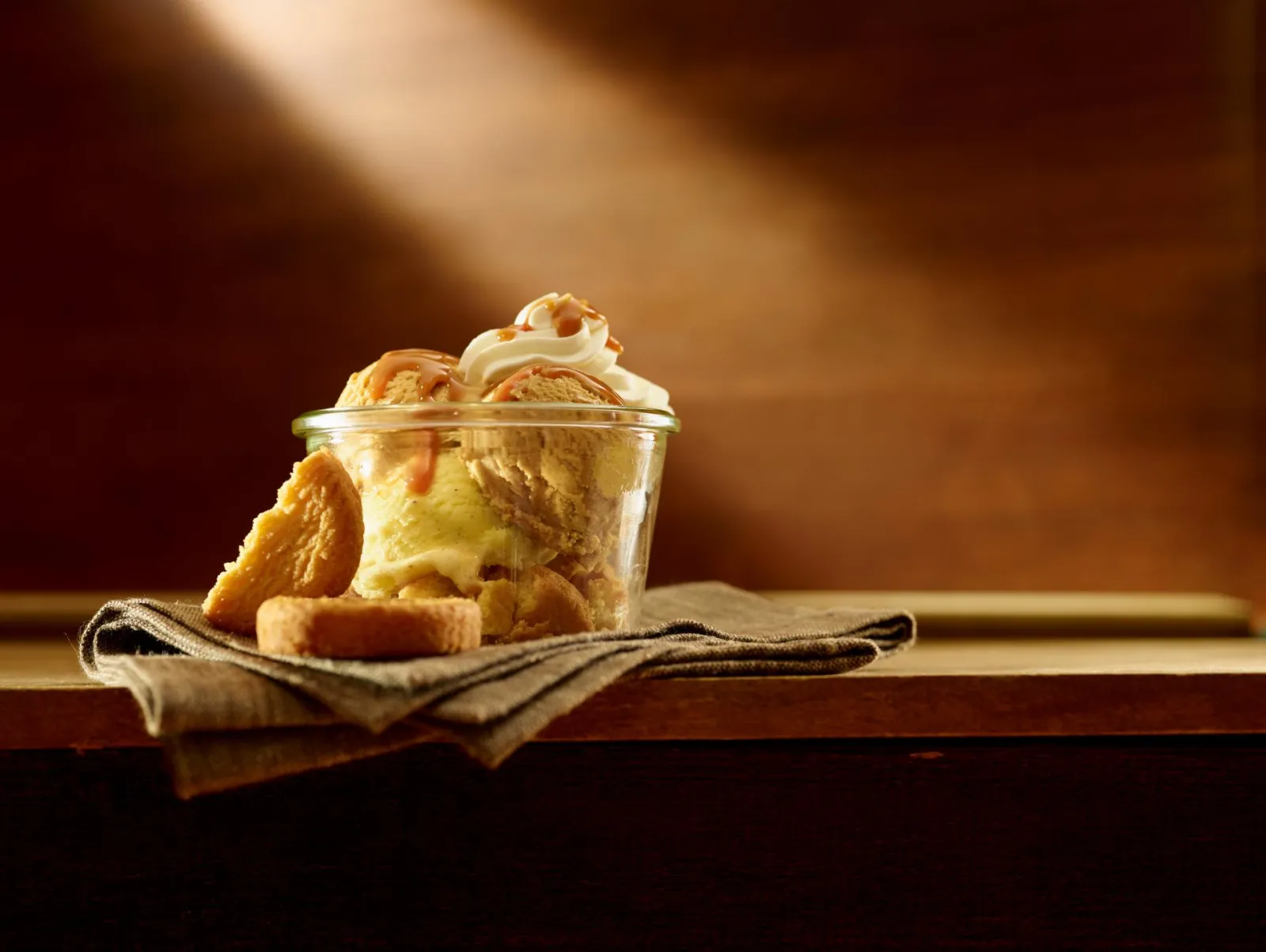 Glass of vanilla ice cream topped with whipped cream and caramel drizzle, served with a cookie on a beige cloth in warm lighting.