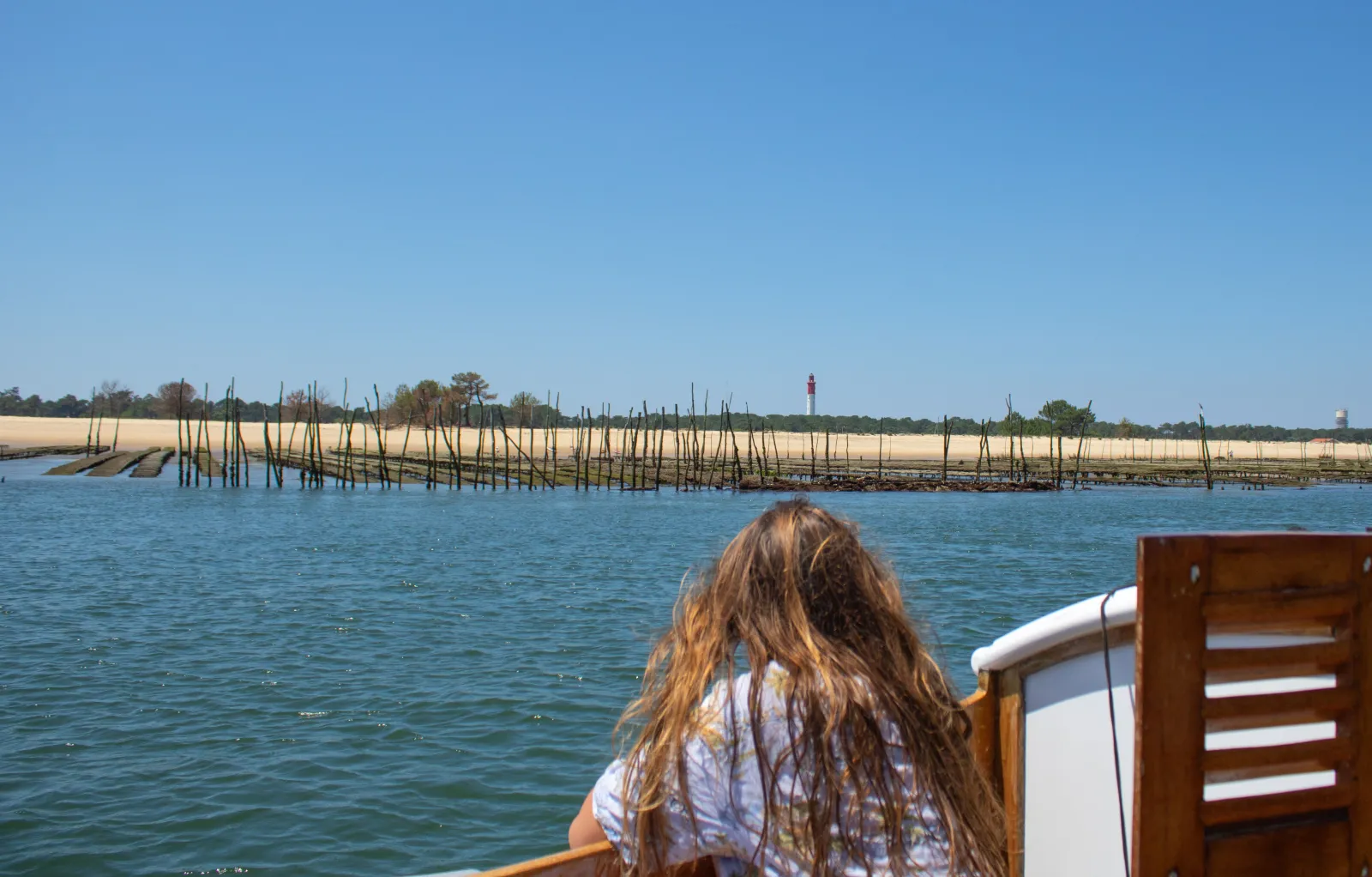 A woman with long hair observes traditional oyster farms from the Pinasse Altaïr on the Gironde estuary near Bordeaux, with a red lighthouse visible on the horizon.