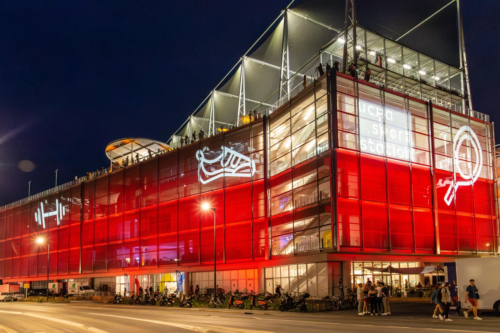 The UCPA Sport Station in Bordeaux lit up at night, a modern building with red façade and glass panels, surrounded by visitors and parked bicycles.