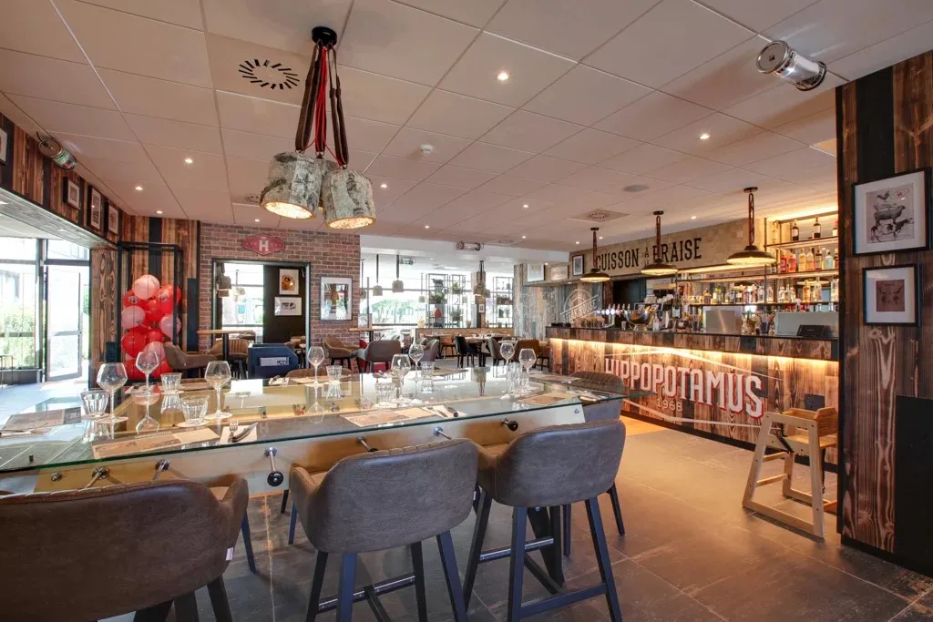 Modern interior of Hippopotamus restaurant in Bordeaux featuring wooden bar counter, high tables, grey chairs, red brick walls, contemporary décor and warm lighting.