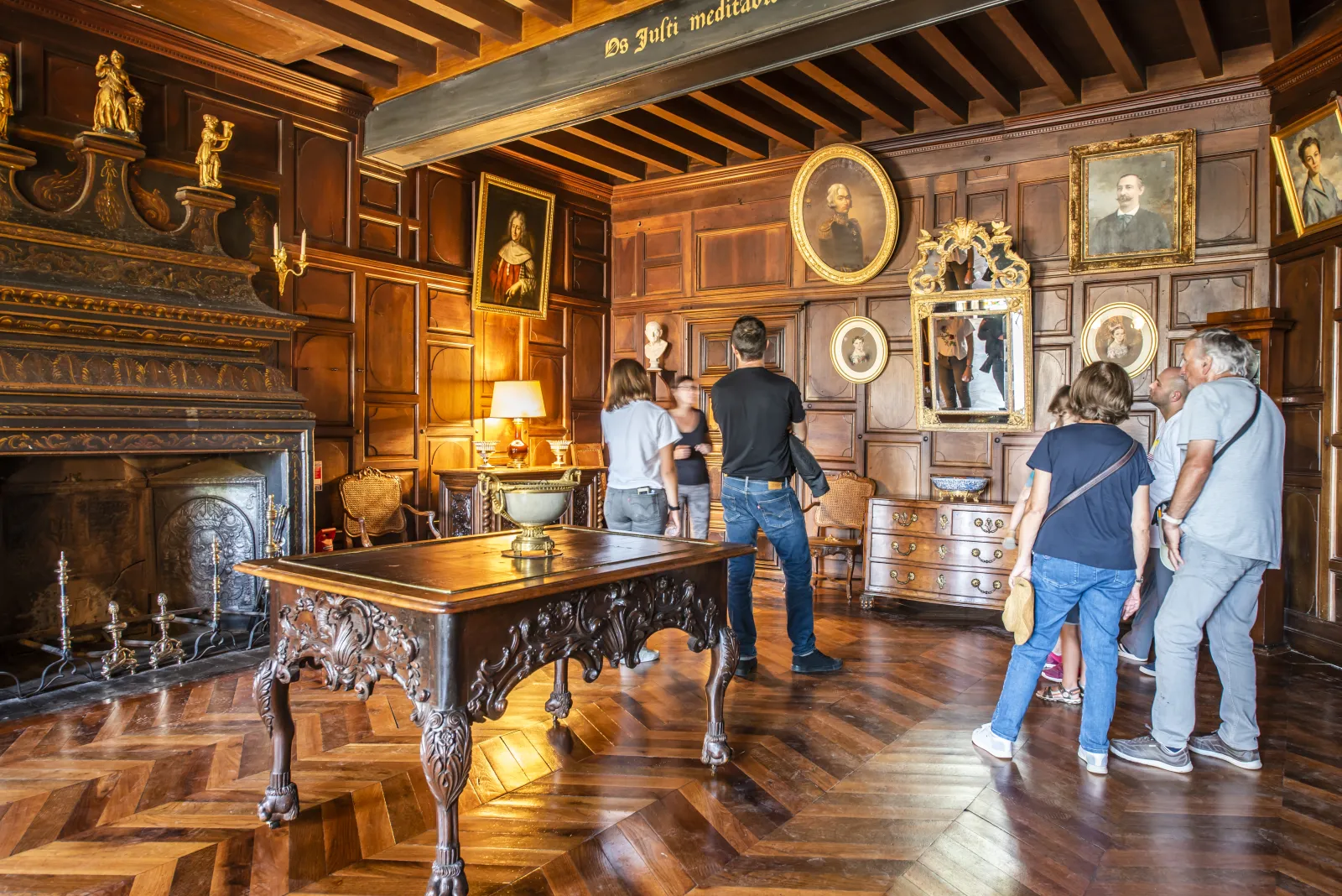 Interior of Château de La Brède near Bordeaux: an ornate room with wood paneling, gilded portrait frames, antique furniture, an elaborate fireplace, and visitors admiring the artworks.