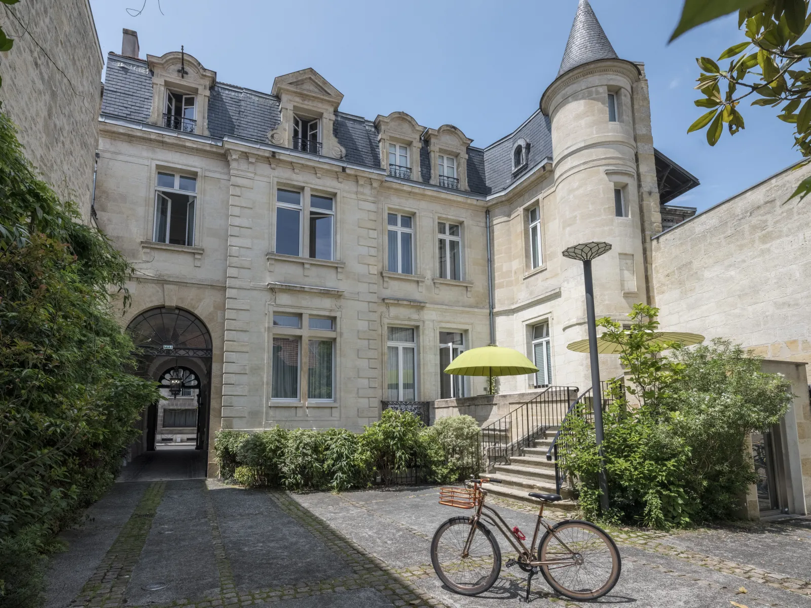 Elegant facade of a Bordeaux mansion hotel with round tower, paved courtyard, parked bicycle, yellow parasol and greenery.
