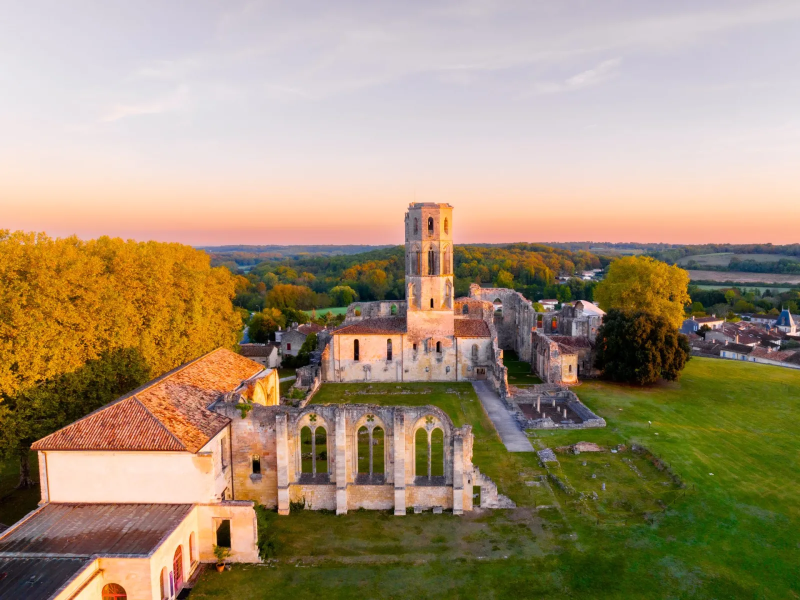 Aerial view at sunset of La Sauve-Majeure Abbey near Bordeaux, showing the church ruins with its Romanesque tower and adjacent buildings surrounded by green fields and forests.