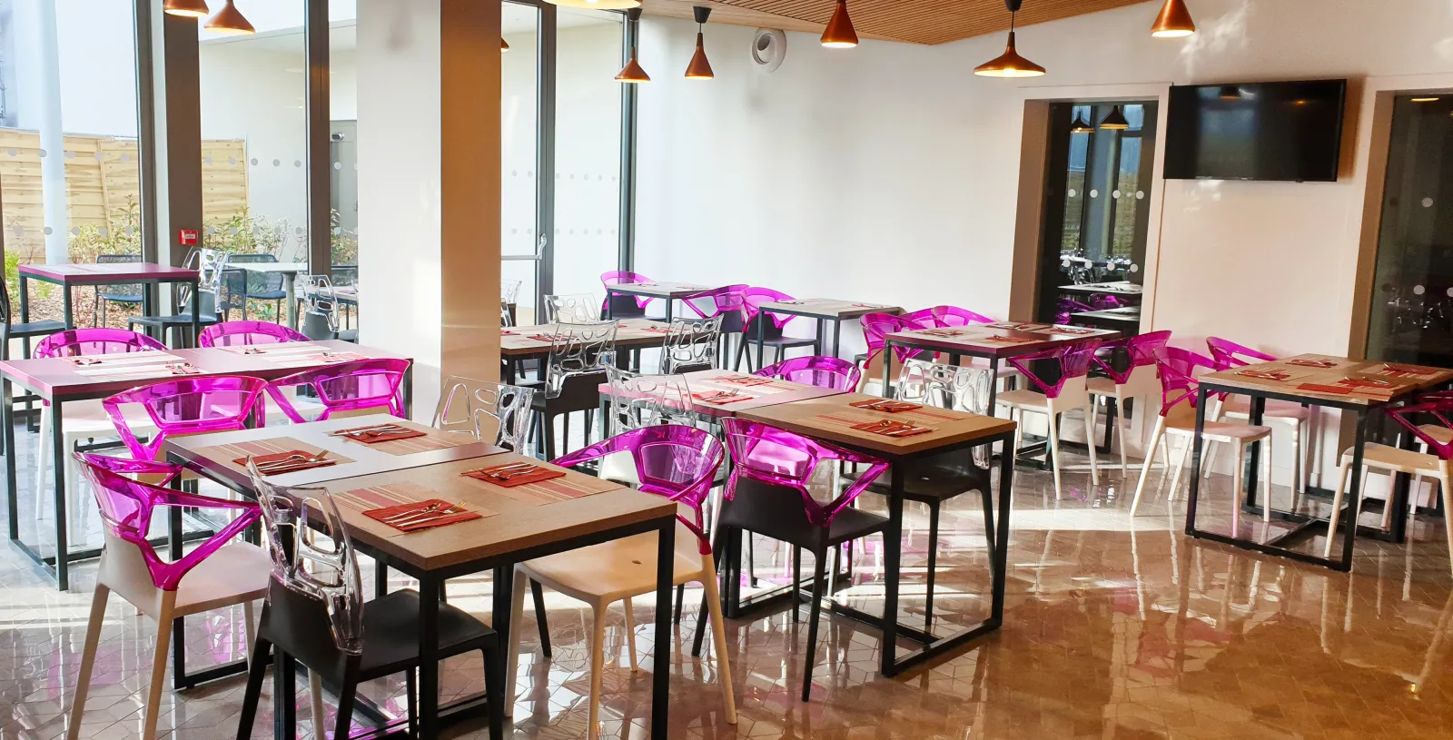 Modern restaurant dining area at Mer & Golf City Apartment Hotel in Bordeaux Lake featuring wooden tables, bright pink transparent chairs, wooden ceiling with copper pendant lights, and floor-to-ceiling windows.