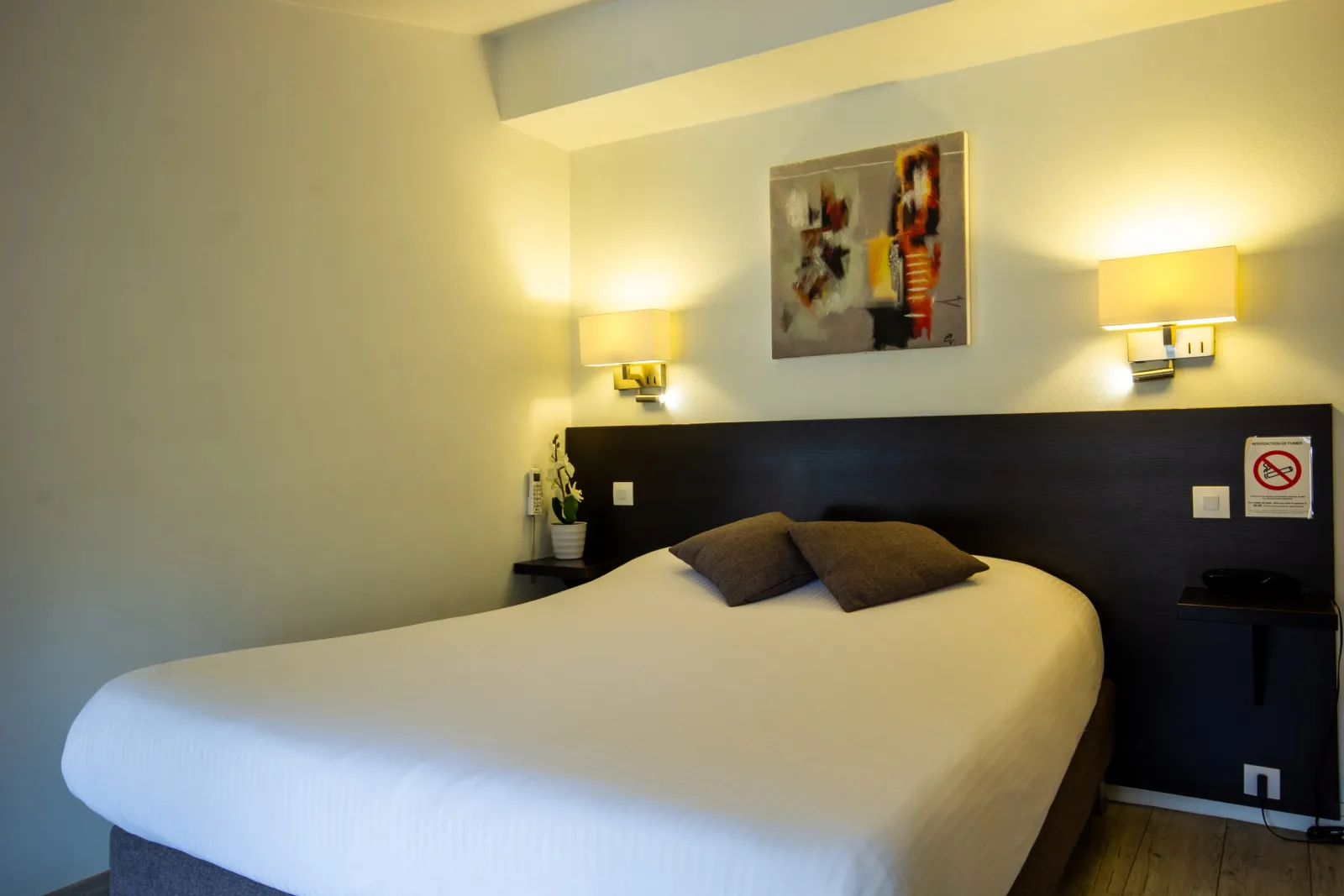 Modern hotel room featuring a white double bed, black headboard, two yellow wall sconces, abstract colorful artwork, and minimalist décor.