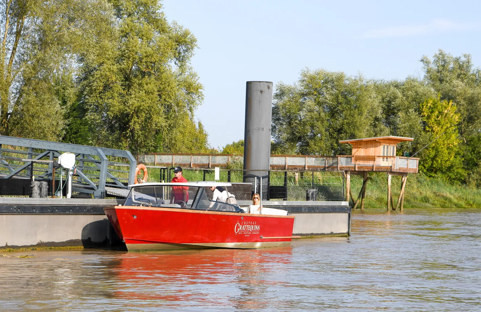 The red Grattequina water taxi carries passengers on the Garonne River in Bordeaux, passing by port facilities and riverside vegetation.