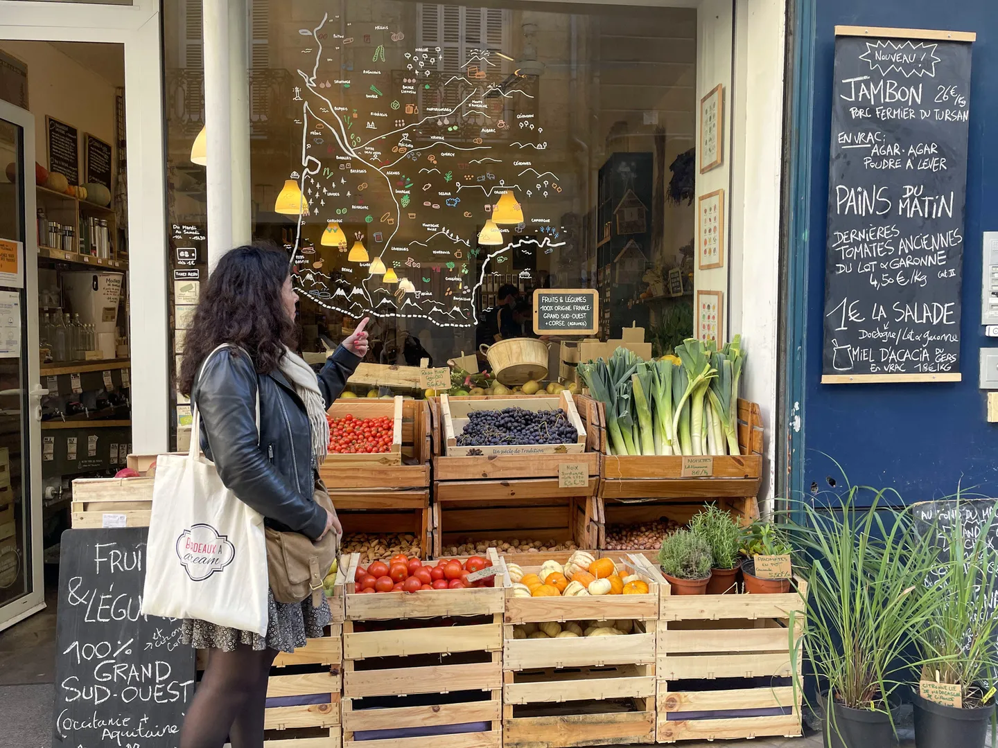 Storefront of a gourmet grocery store in Bordeaux displaying fresh produce in wooden crates, a hand-drawn map of France on the window, and daily menu on a blue chalkboard.