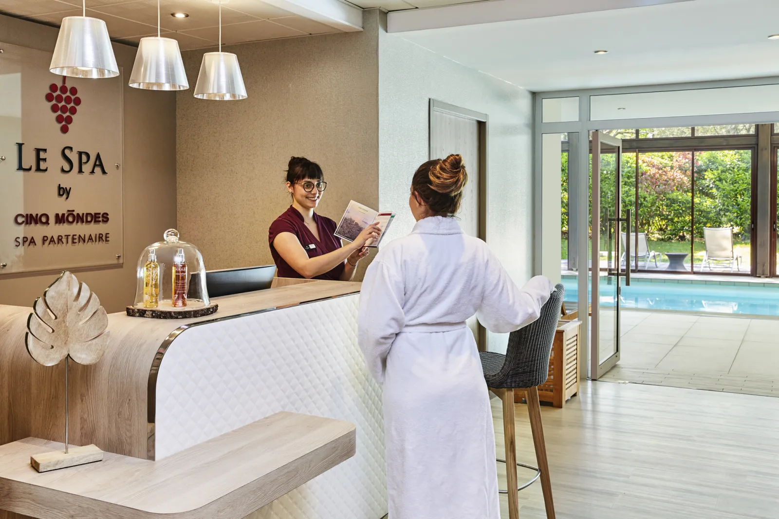 Reception desk at Le Spa by Cinq Mondes at Cabot Bordeaux MGallery Collection hotel. A spa receptionist in burgundy uniform greets a guest in a white robe. Modern reception area overlooks the indoor swimming pool through large windows.
