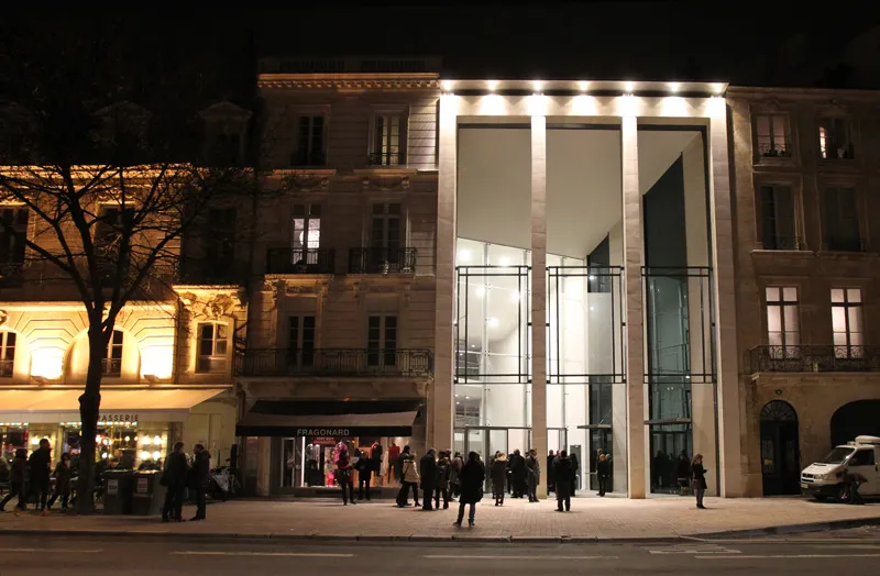 Night view of the Bordeaux Auditorium with its modern illuminated glass façade, surrounded by historic Haussmann-style buildings, with visitors gathered at the entrance plaza.