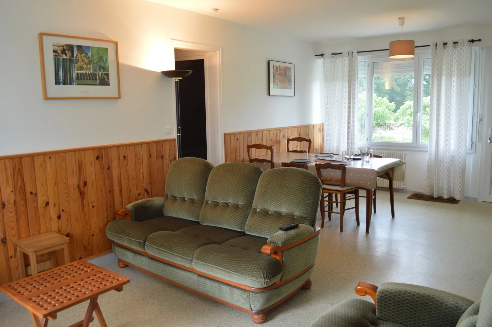 Spacious living room in a cottage featuring an olive-green sofa, wooden chairs, wooden wall paneling, dining table, large windows overlooking a garden, and warm rustic decor.