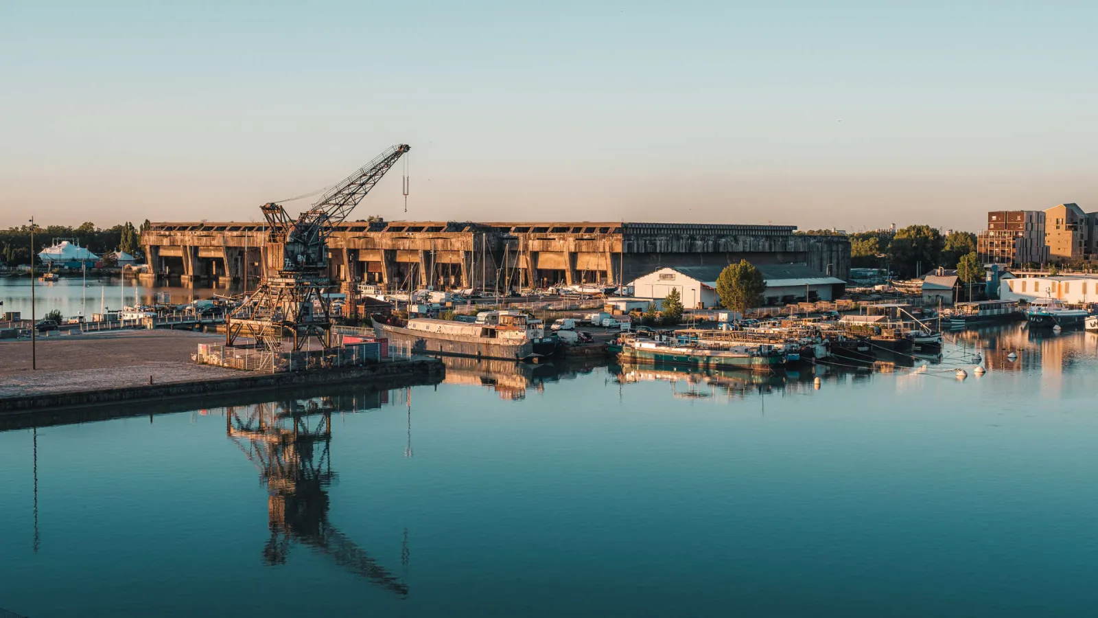 Sunset view of Bordeaux's submarine base featuring concrete bunkers, a port crane, and moored boats reflected in the calm waters of the Garonne River.