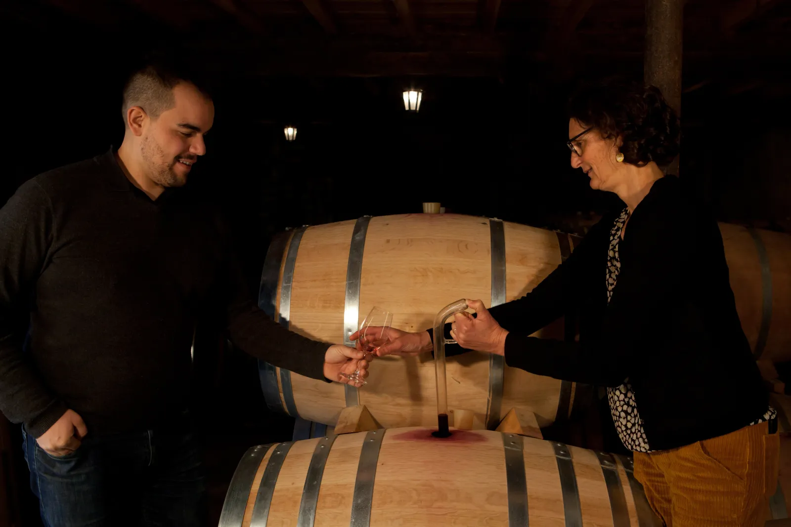 Two people toasting with glasses of red wine in front of wooden barrels in a Bordeaux wine cellar.