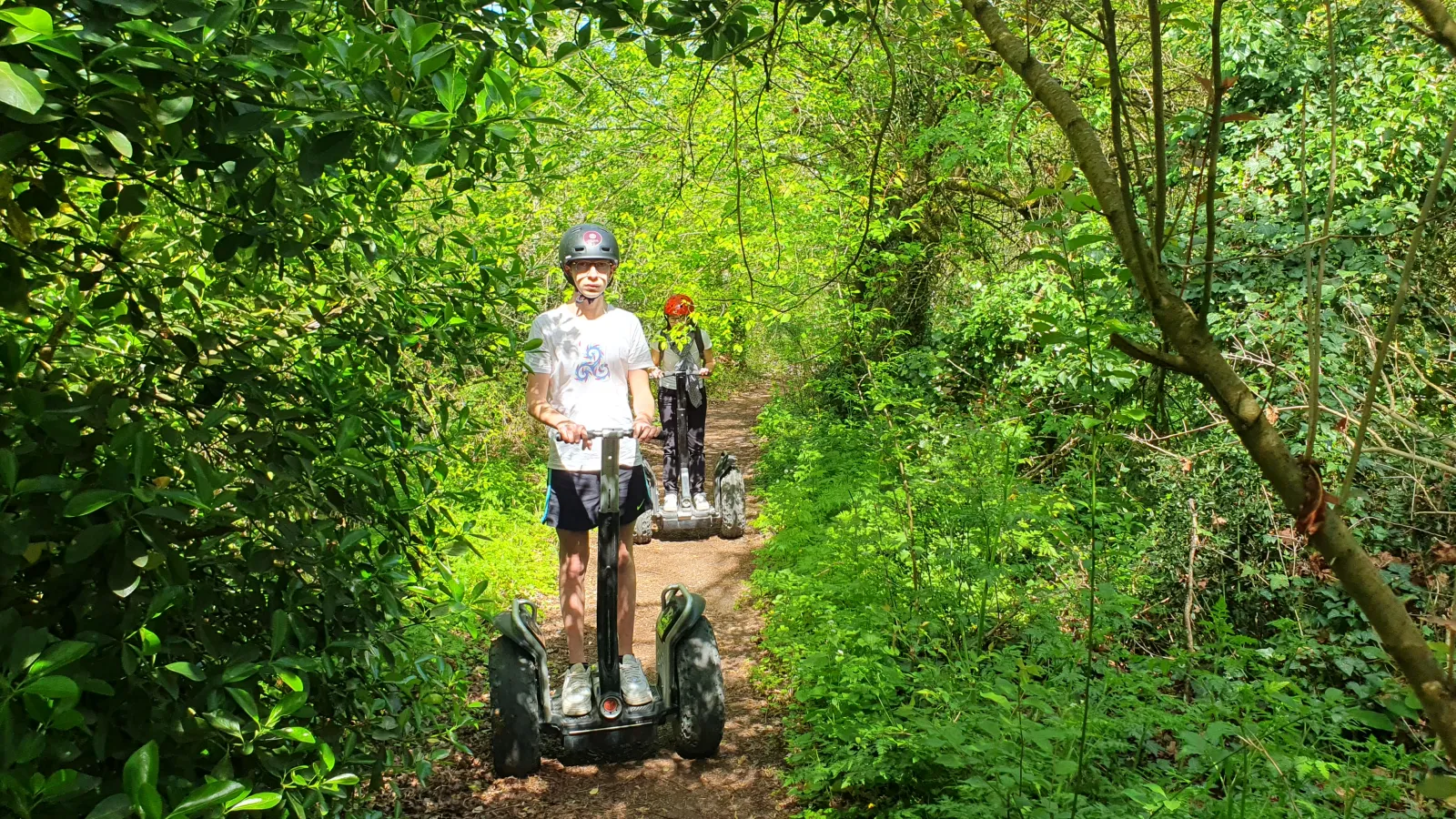 Two people riding Segways on a shaded forest trail wearing protective helmets during a guided tour in Pessac-Léognan near Bordeaux.