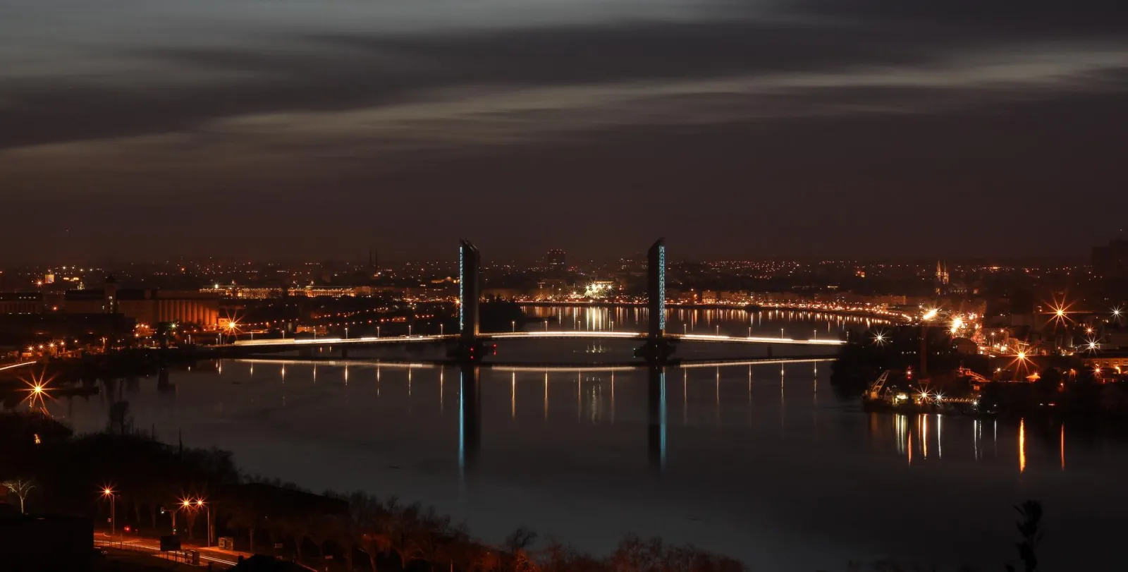 Night view of the Jacques Chaban Delmas Bridge in Bordeaux, lit up and reflected in the Garonne River, with the sparkling city in the background