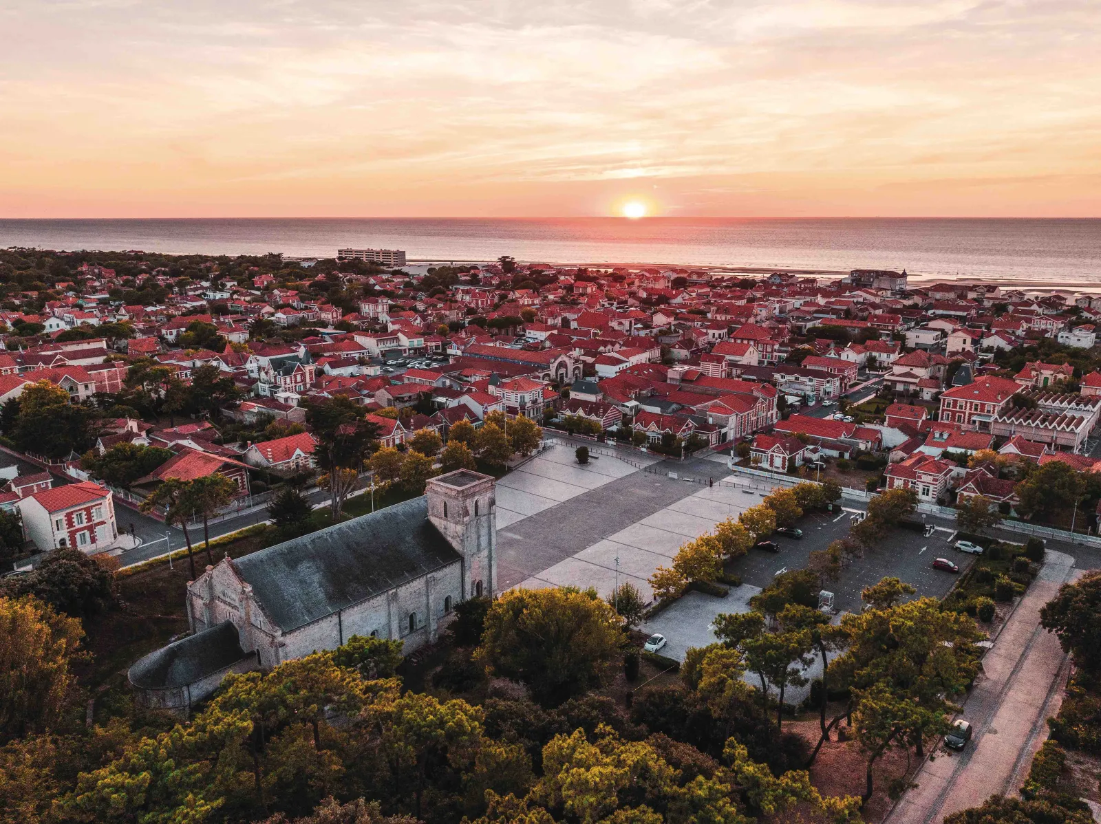 Aerial sunset view of the Notre-Dame-de-la-fin-des-Terres Basilica in Bordeaux, surrounded by red-roofed houses with the Atlantic coast in the background.
