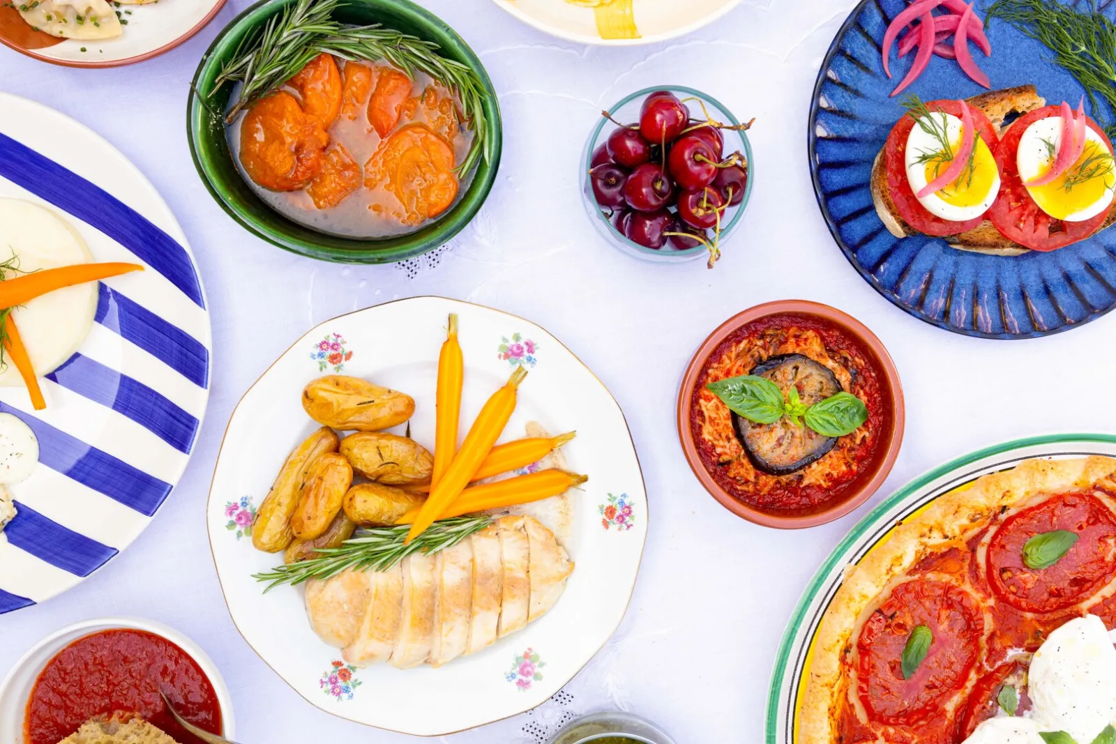 Colorful kitchen table featuring a variety of Mediterranean dishes including grilled fish, vegetables, fruits, topped toast, and sauces served on decorated plates with blue striped and floral patterned tableware.