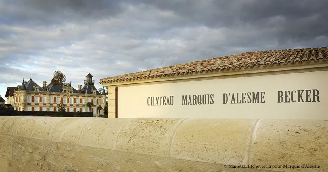 Entrance wall of Château Marquis d'Alesme Becker in Bordeaux displaying the château's name, with the historic building visible in the background under a cloudy sky.