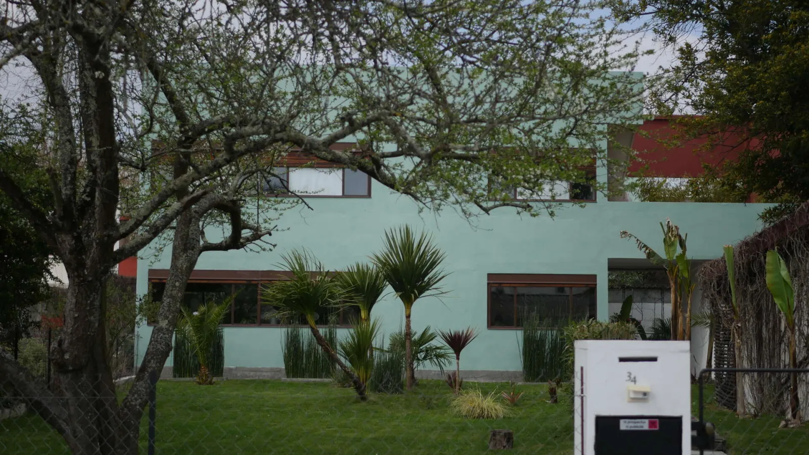 Modernist house in Cité Frugès, Pessac, featuring pale green painted facade, rectangular windows, flat roof, and landscaped garden with palm trees and mature trees.