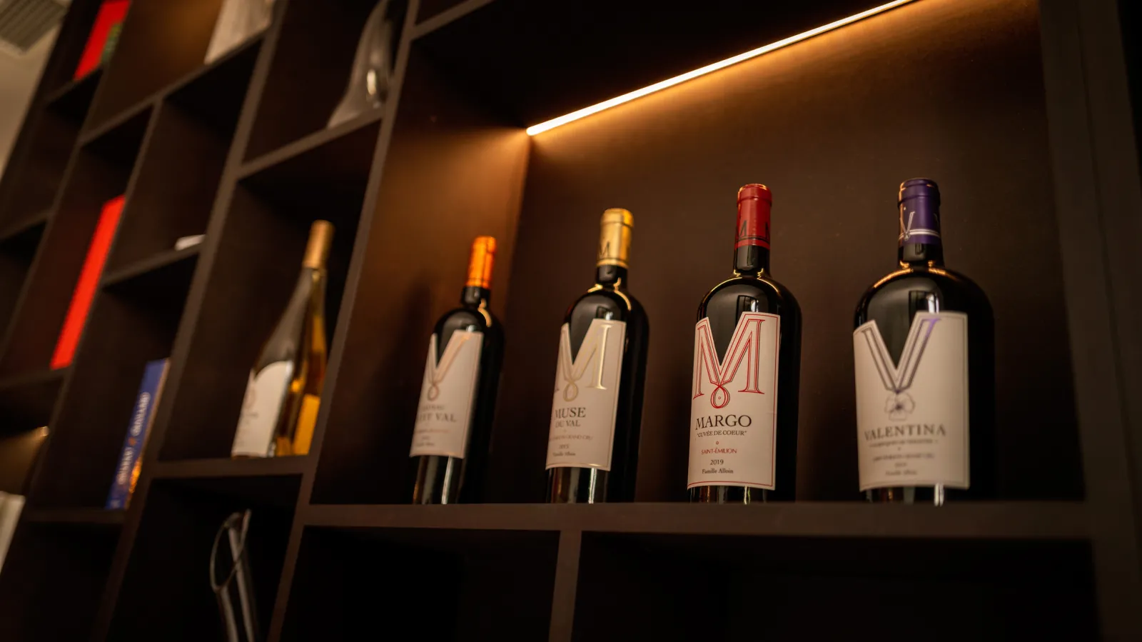 Display of red wine bottles from Château Petit Val arranged on a lit shelf, featuring Muse, Margo, and Valentina vintages in a Bordeaux wine cellar.