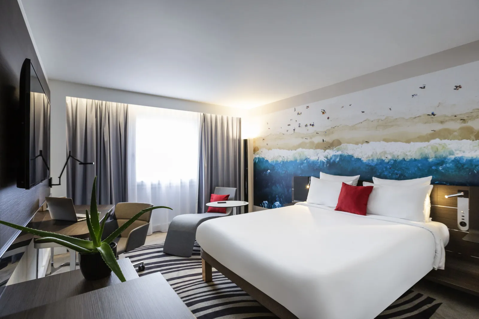 Modern hotel room at Novotel Bordeaux Centre featuring a queen bed with white linens, blue forest panoramic wall art, workspace with desk, lounge chair, wall-mounted TV, and floor-to-ceiling windows