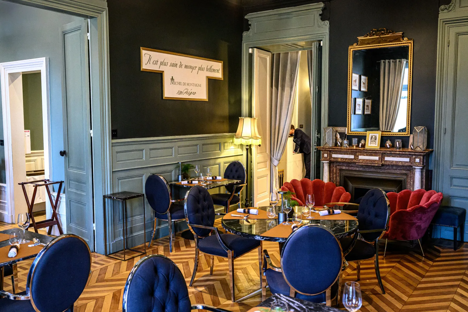 Elegant room at Cercle de Montaigne in Bordeaux featuring blue and red chairs, black tables, herringbone flooring, marble fireplace, gilded mirror, and framed French quote on dark wall.