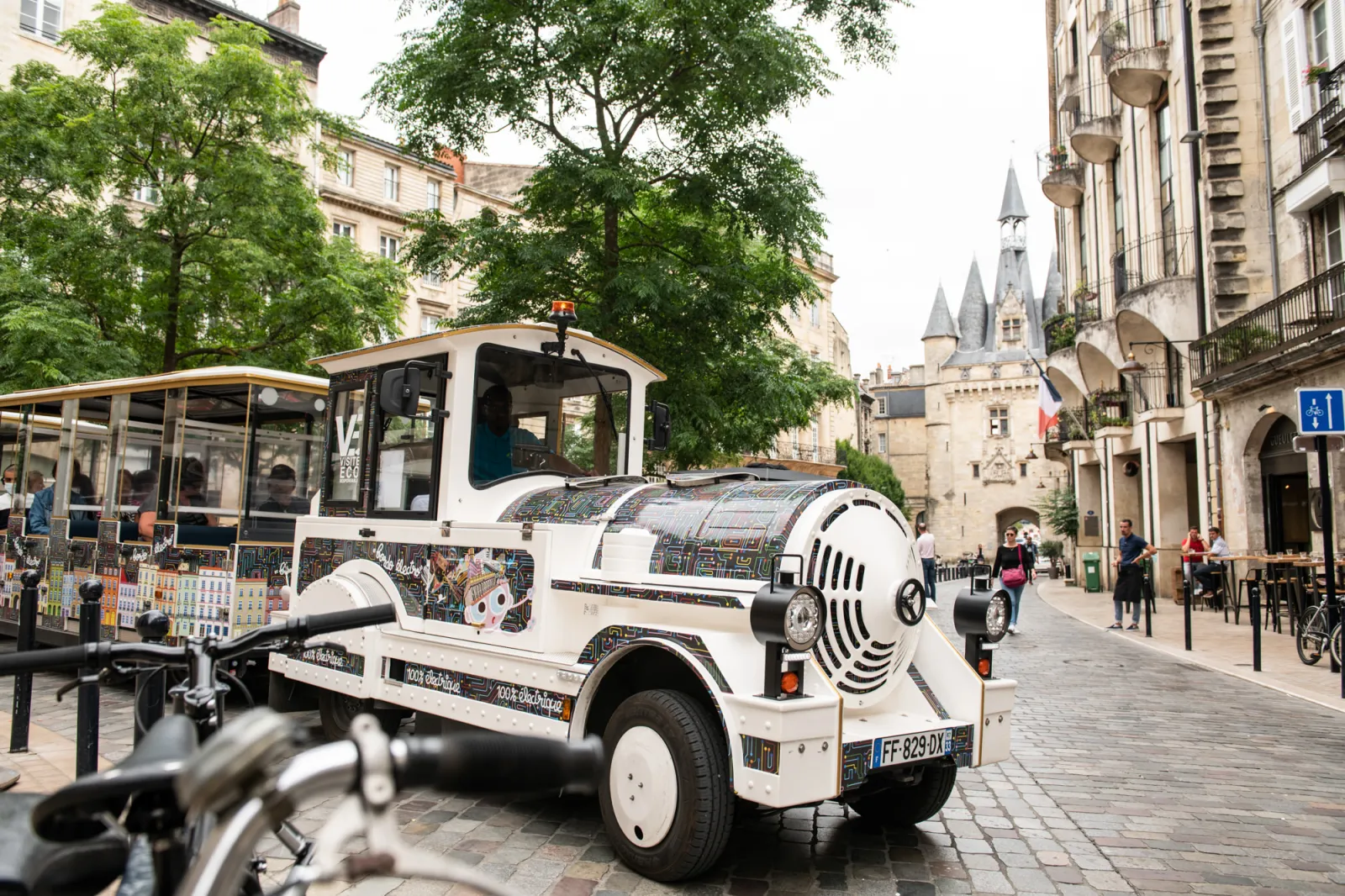 A white and black electric tourist train traveling down a cobblestone street in Bordeaux, with Haussmann-style buildings and a castle in the background.
