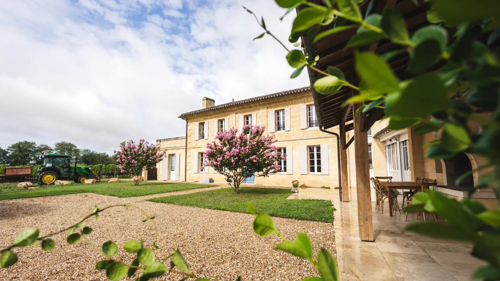 Château La Voûte near Bordeaux: stone-built mansion with yellow ashlar facade decorated with pink flowering shrubs, gravel driveway, covered terrace, and farm tractor in the surrounding countryside.