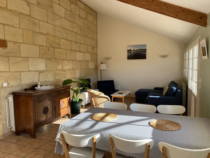 Common room of the Dovecote at Château La Gontrie: spacious living area with gray patterned dining table, natural wood chairs, antique wooden furniture, green plant, exposed stone walls, wooden beams, black sofa and glass doors opening to the outdoors.