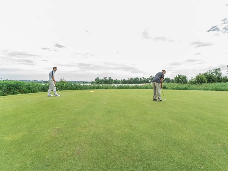 Two golfers on the green at Golf de Margaux near Bordeaux, with lush landscape and cloudy sky in the background.