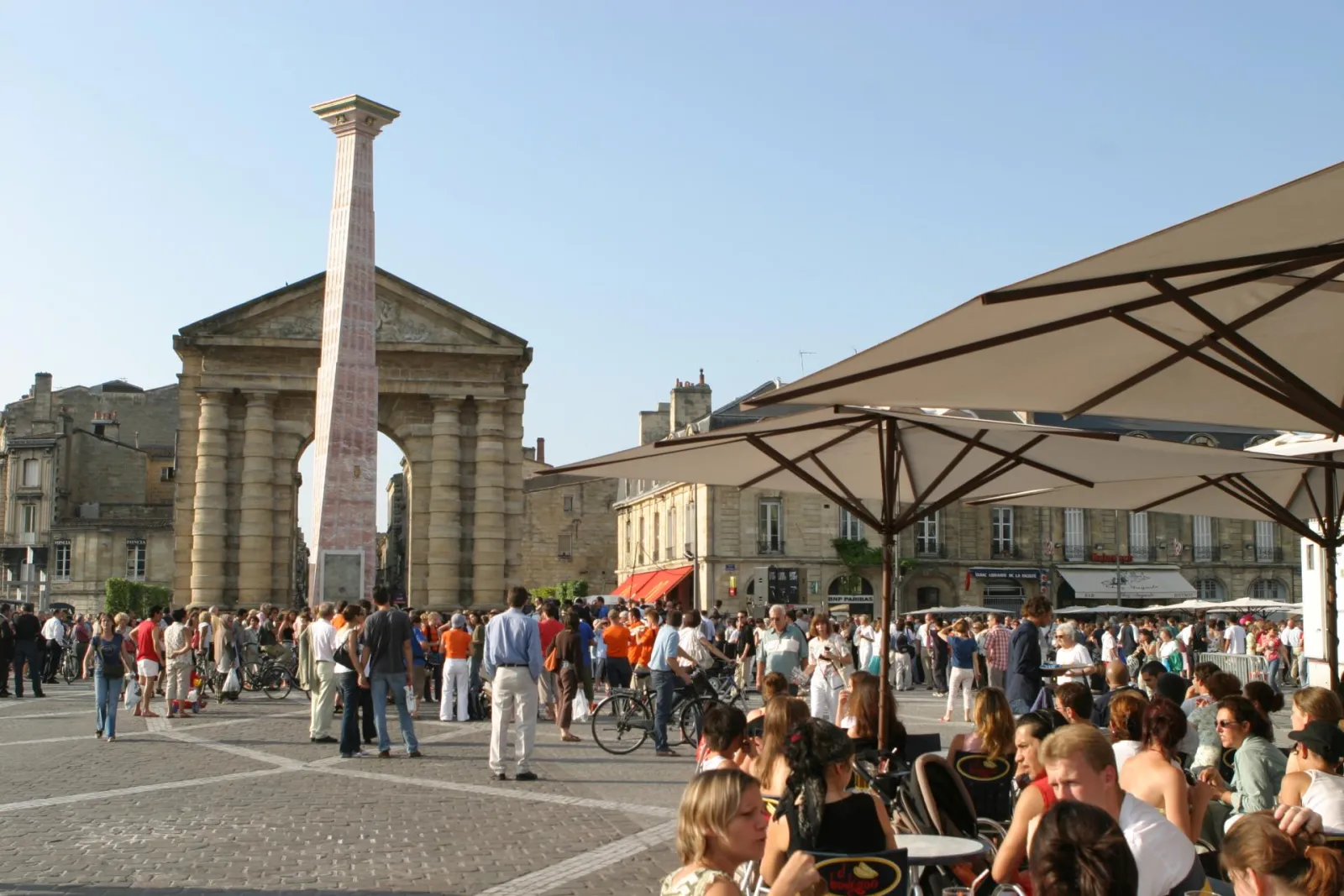 Victory Square in Bordeaux featuring the commemorative column, a church, outdoor café umbrellas, and crowds of visitors on a sunny day.