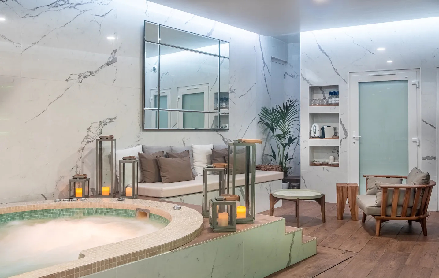 Luxury spa space featuring a circular thermal pool with green tile trim, elegant wood and linen furniture, white marble walls, modern mirror, plants and ambient candlelight