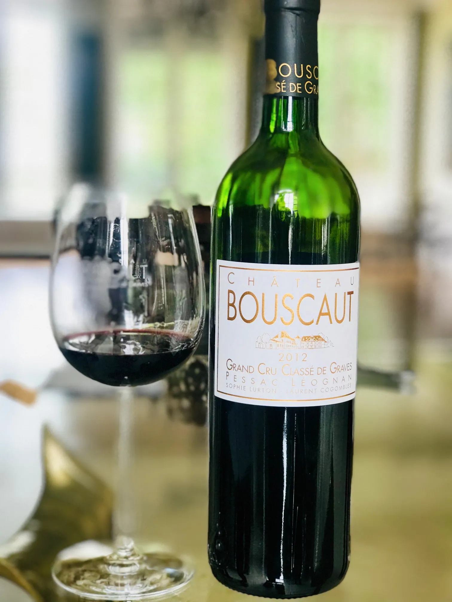 Bottle of red wine Château Bouscaut, Grand Cru Classé de Graves, with a glass of poured wine against a blurred indoor background.