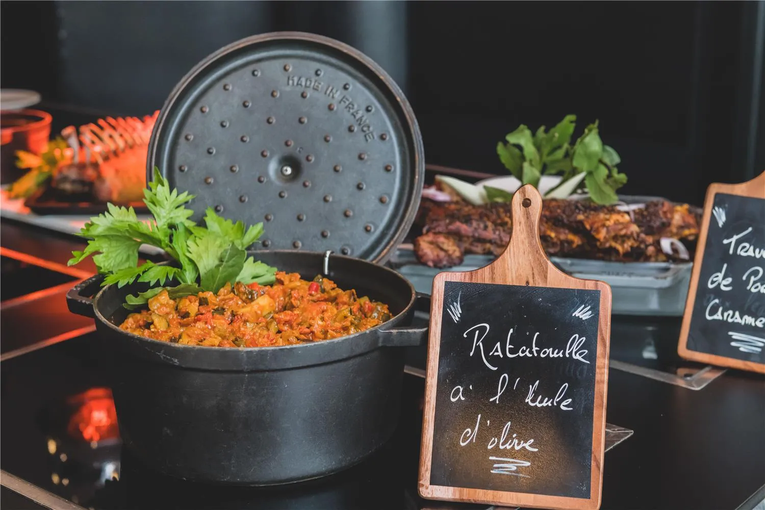 A black pot with lid containing ratatouille garnished with fresh parsley, accompanied by a wooden chalkboard menu sign reading 'Ratatouille à l'huile d'olive', displayed in a Campanile restaurant in Bordeaux