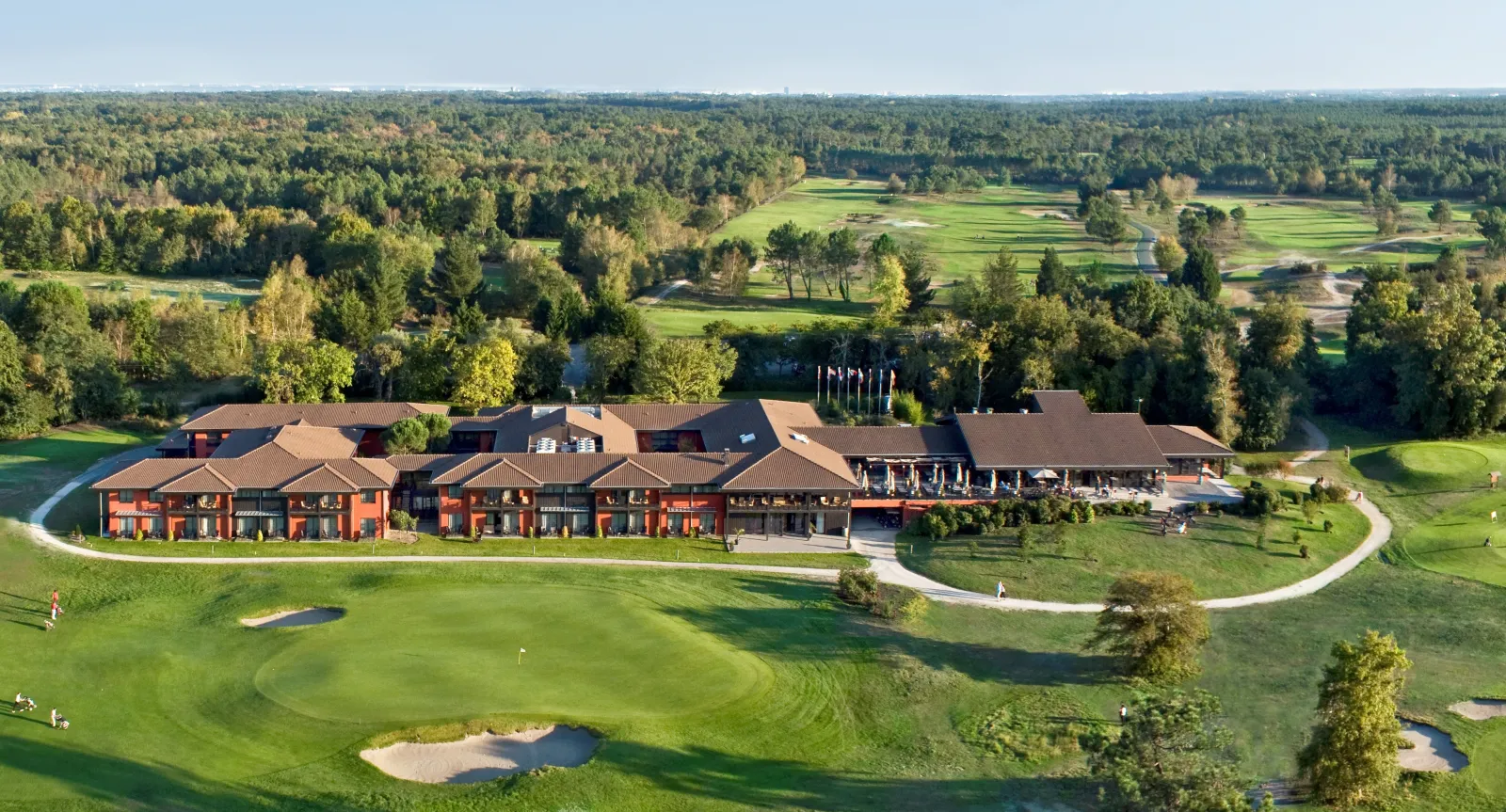 Aerial view of a luxury hotel complex with red-orange facades and brown roofs, surrounded by a lush golf course with bunkers, water features, and forests near Bordeaux.