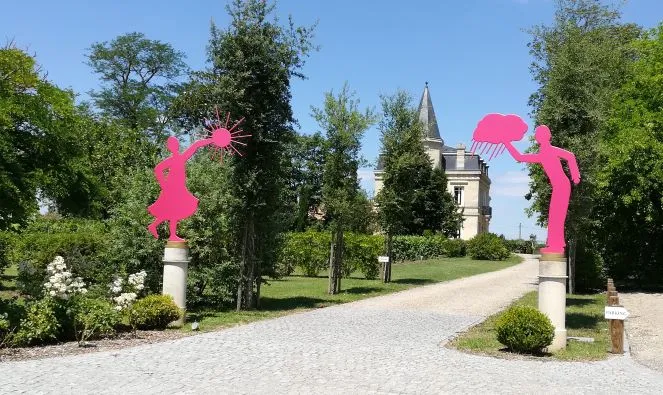 Driveway leading to Château Yon-Figeac in Bordeaux, lined with bright pink abstract figure sculptures on both sides, surrounded by green trees with the castle tower visible in the distance.