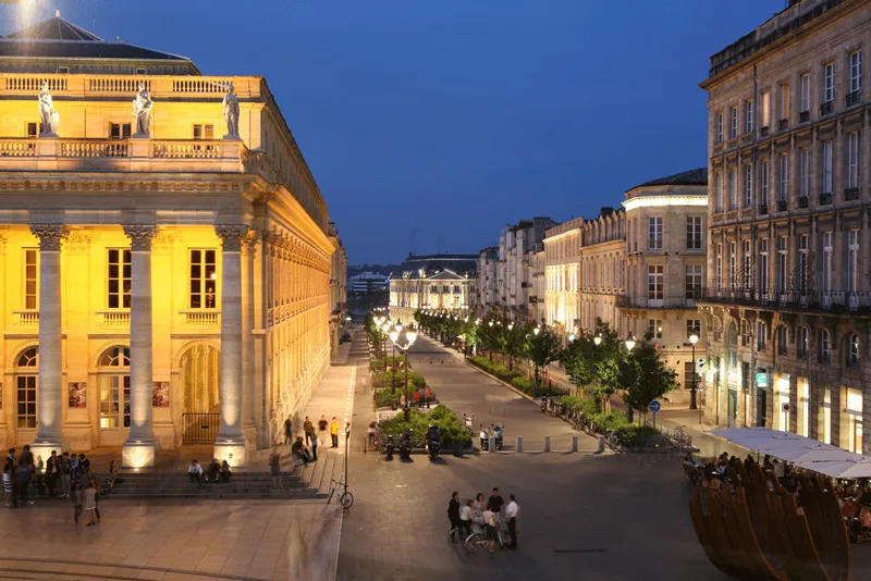 Night view of Bordeaux's Grand Theatre brightly lit in yellow, featuring its impressive columns and neoclassical facade, overlooking a lively pedestrian square surrounded by Haussmann-style buildings.