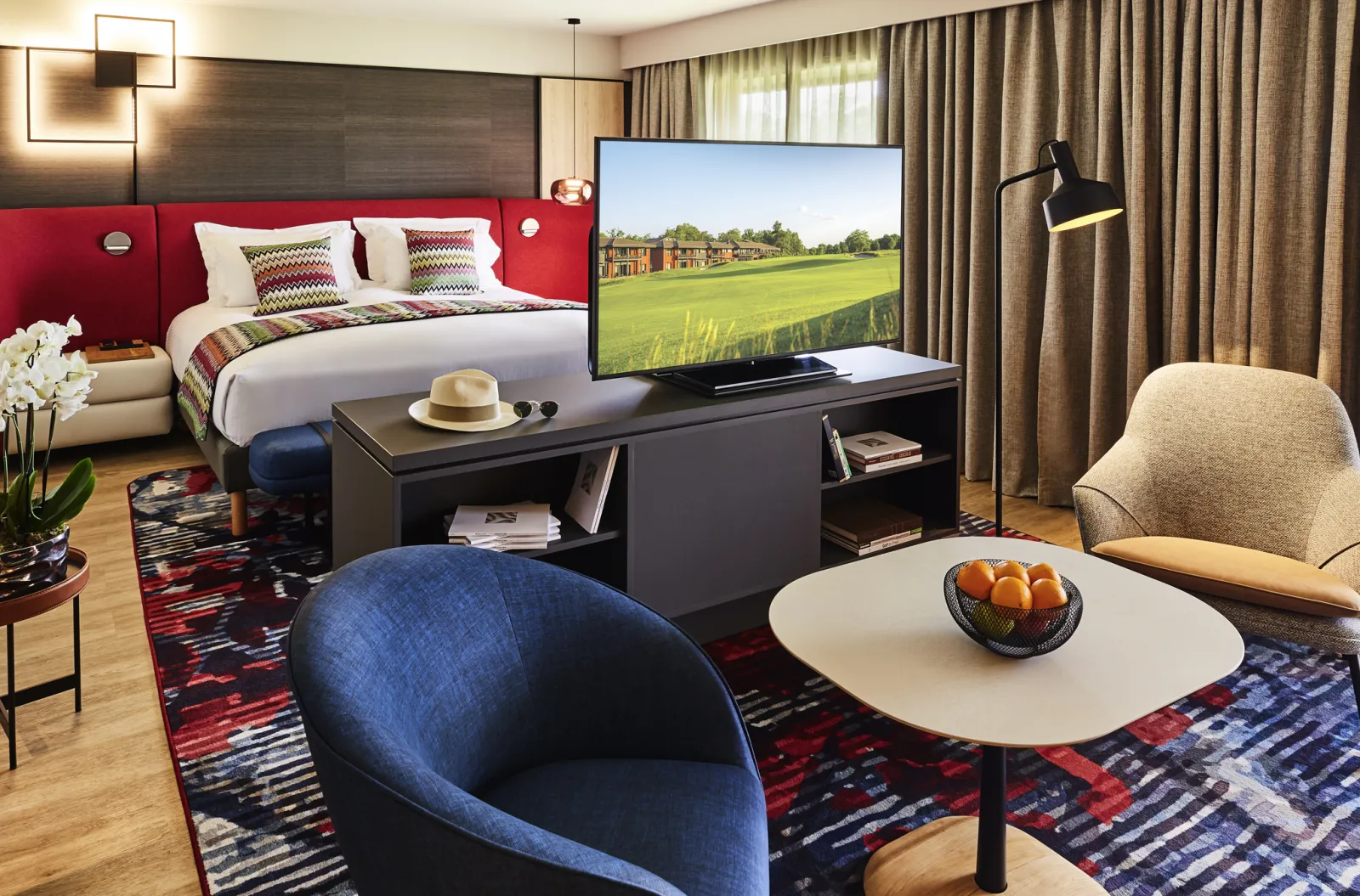 Luxury hotel suite featuring a red upholstered bed, sitting area with blue and beige armchairs, television displaying green landscape, beige curtains and contemporary décor.