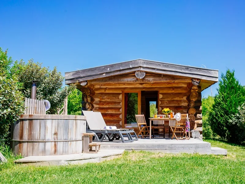 Wooden log cabin with furnished deck, outdoor hot tub, and garden furniture surrounded by lush greenery at Domaine EcÔtelia in Bordeaux.
